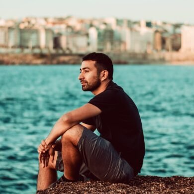 man in purple shirt and gray pants sitting on brown rock near body of water during