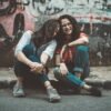 two women sitting on pavement near painted wall